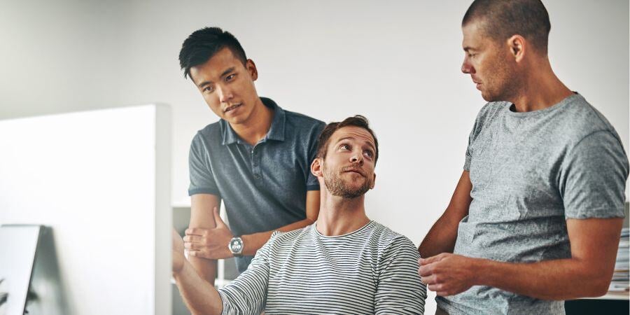 3 Colleagues Looking At Computer Screen At Coworkers Desk