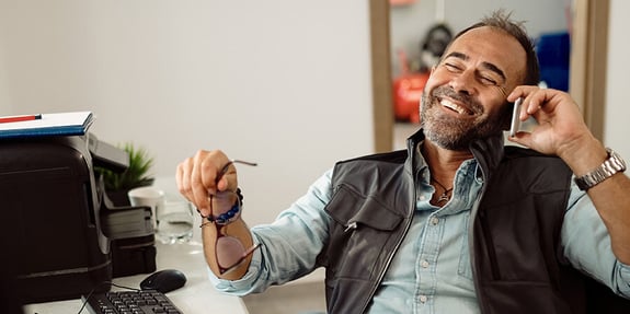 Happy man holding his glasses while talking on the phone at his desk.