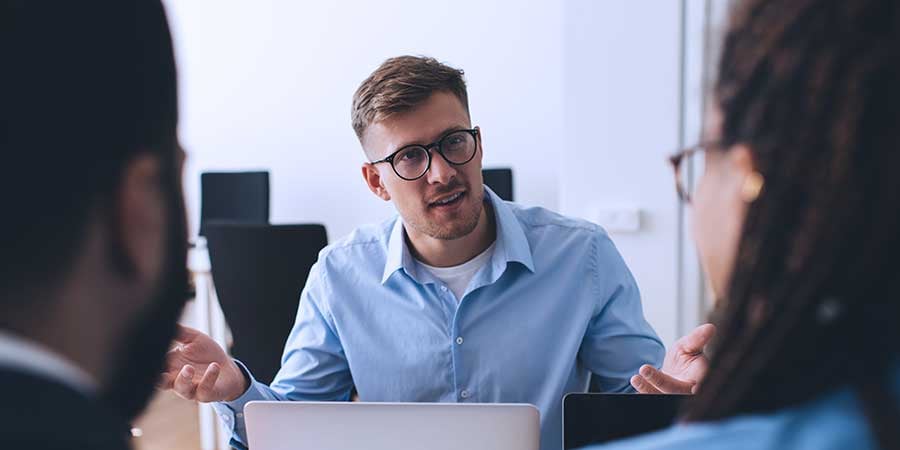 Man wearing glasses talking to two team members with computer at table