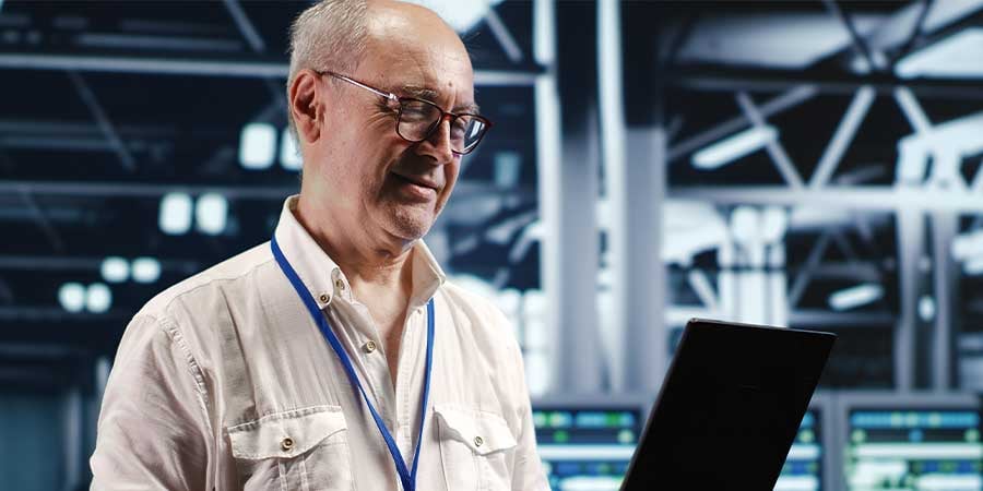 A man wearing glasses holding a laptop in his hands while standing in a data warehouse