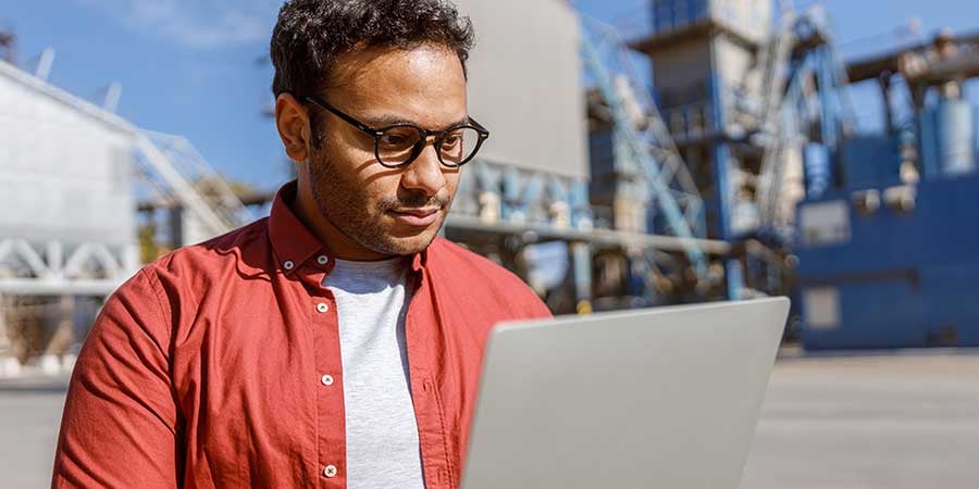 Man with glasses looking at their computer while holding it on an industrial lot. 