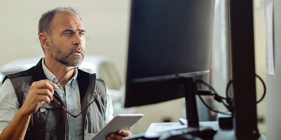 Worried Man Looking At Tablet And Computer In Warehouse